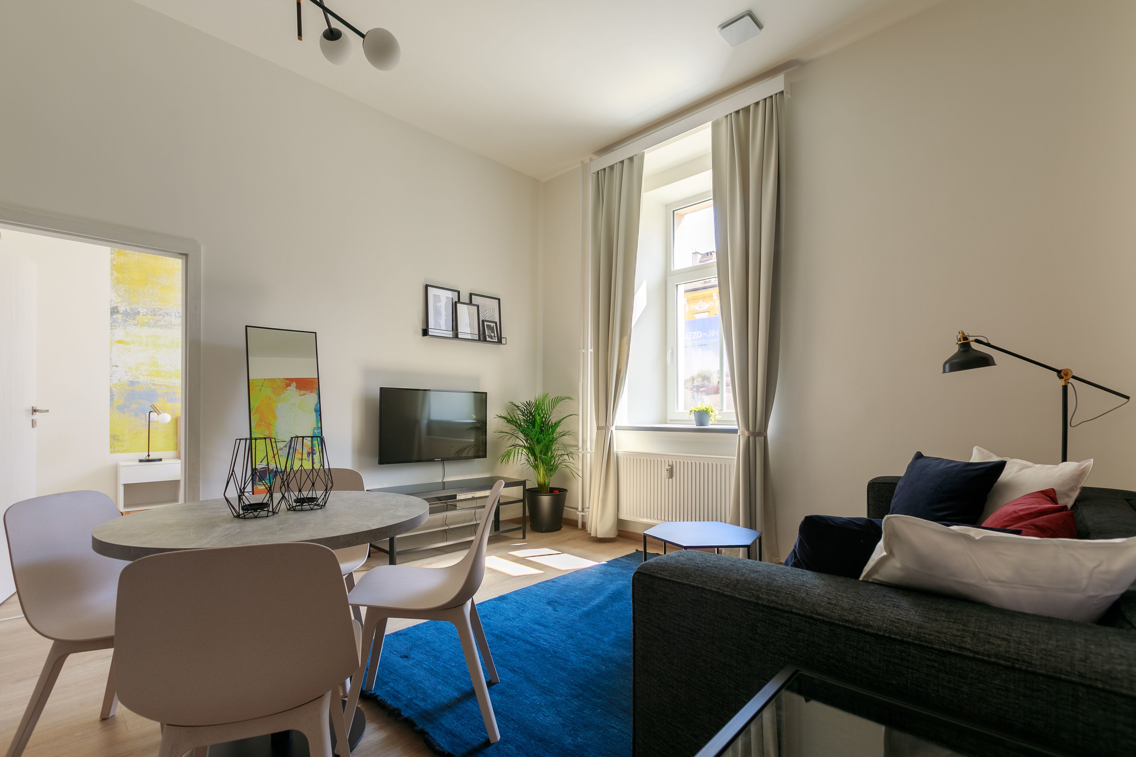 A modern living room with a sofa, a dining table with chairs, a TV, and a window with curtains.