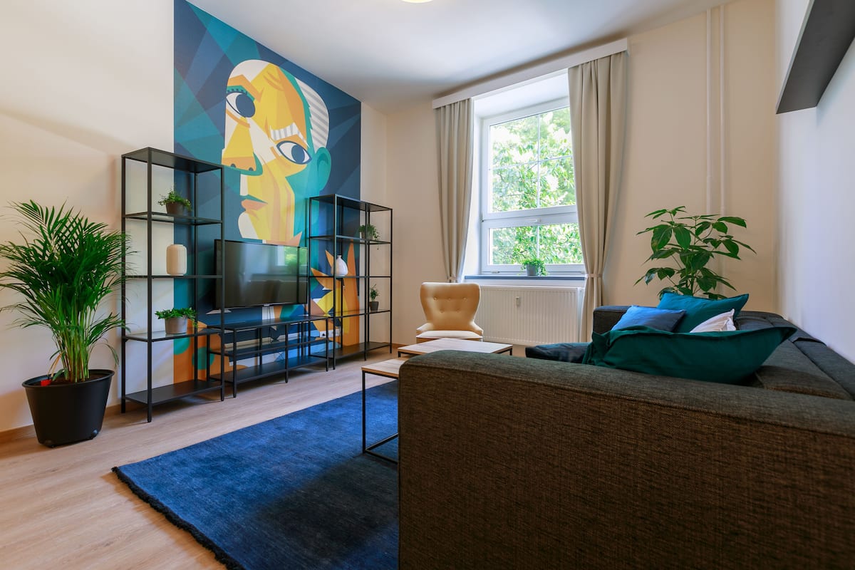 A modern living room with a large abstract painting, a sofa, a chair, a TV on a black stand, and a potted plant.