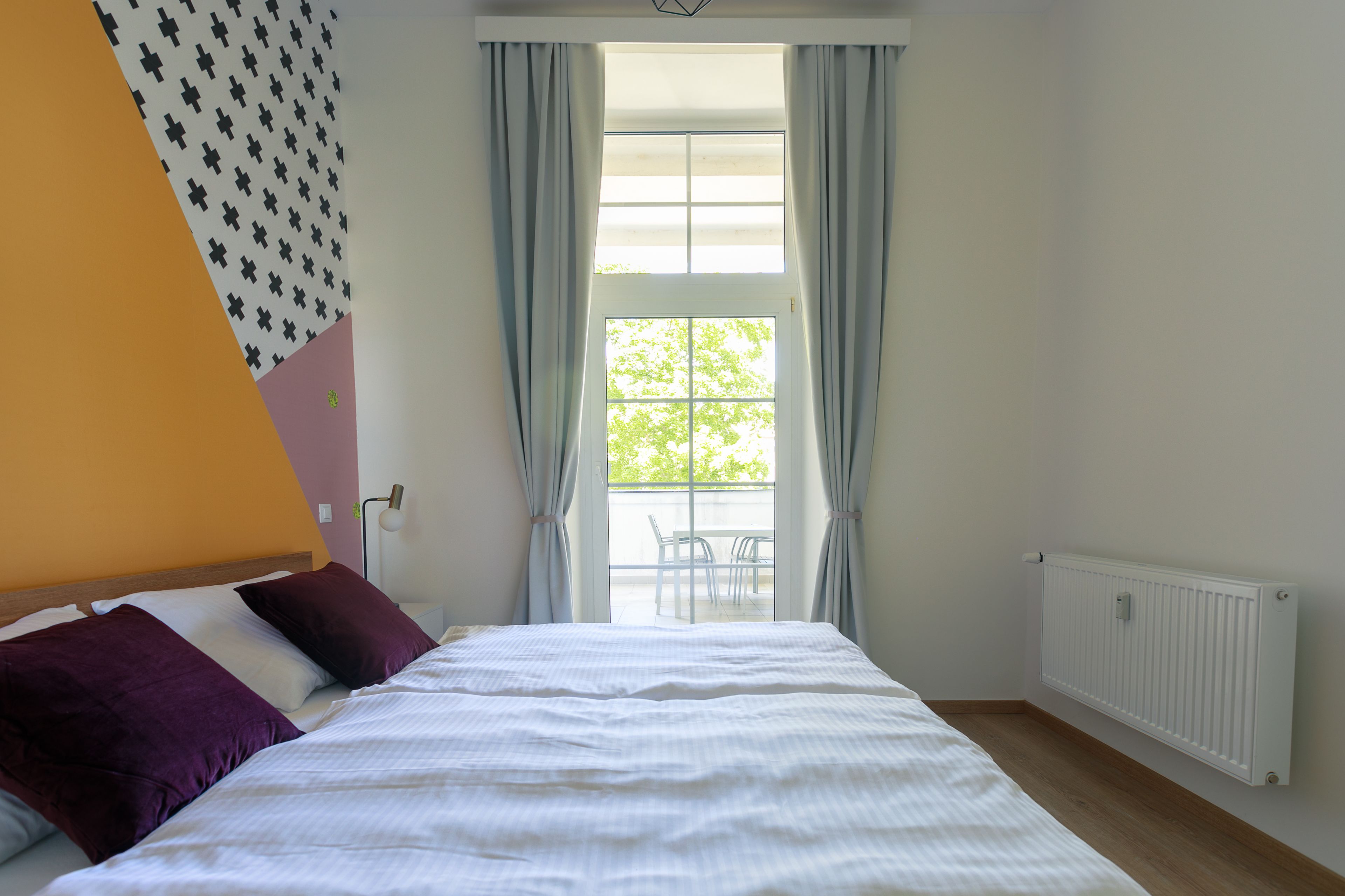 Ein Schlafzimmer mit Bett, einem Fenster mit Vorhängen, einem Heizkörper und einem Balkon mit Stühlen.