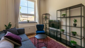 A modern living room with a grey sofa, a blue armchair, a black TV stand, and a black shelving unit with potted plants. - Avantgarde apartments (Plzen)