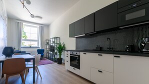 A modern kitchen with white cabinets, a black countertop, and a built-in oven. There is a dining area with a table and chairs, and a potted plant. - Avantgarde apartments (Plzen)