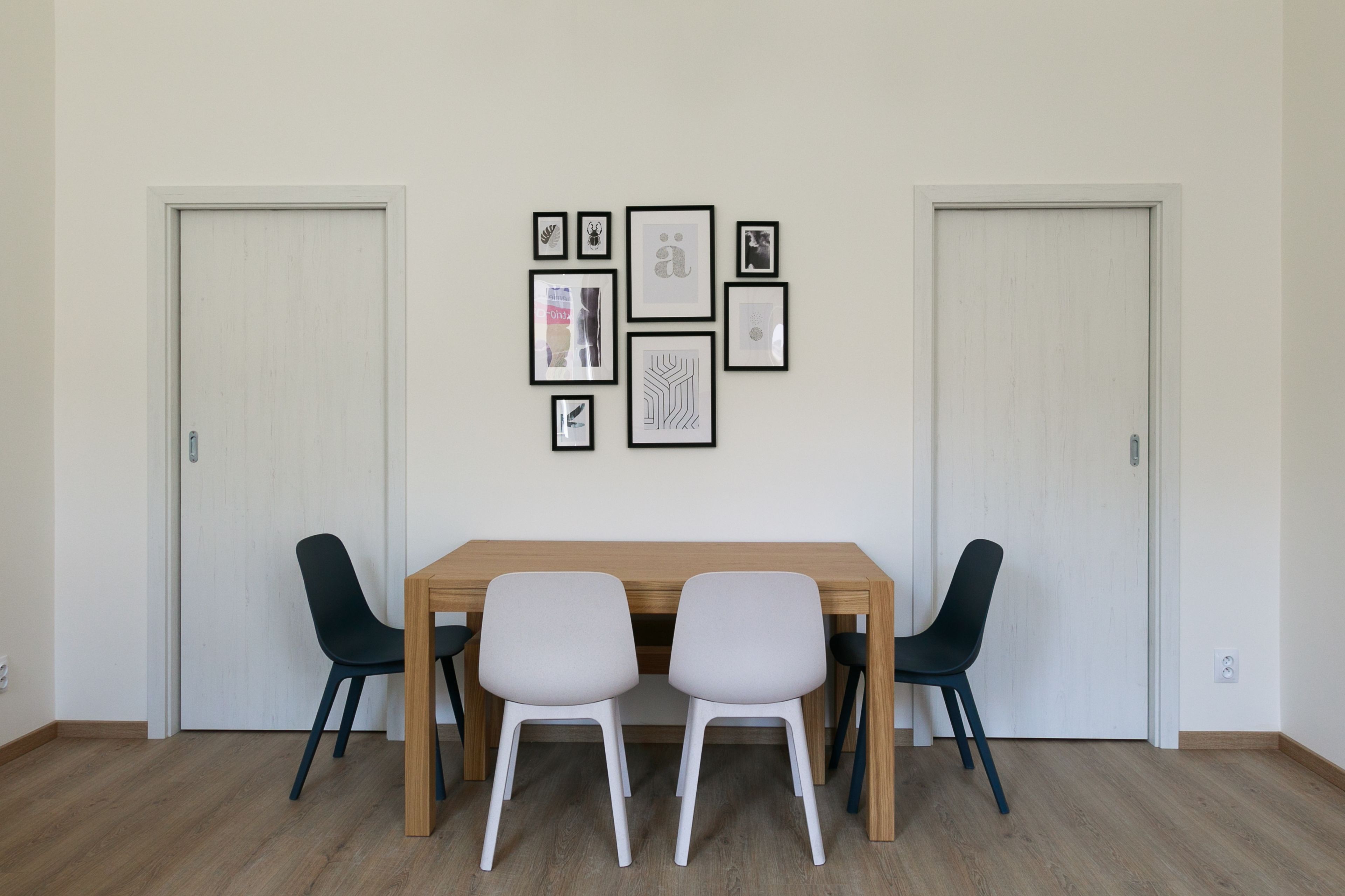 Un coin repas avec une table en bois et deux chaises, des œuvres d’art encadrées au mur et deux portes.