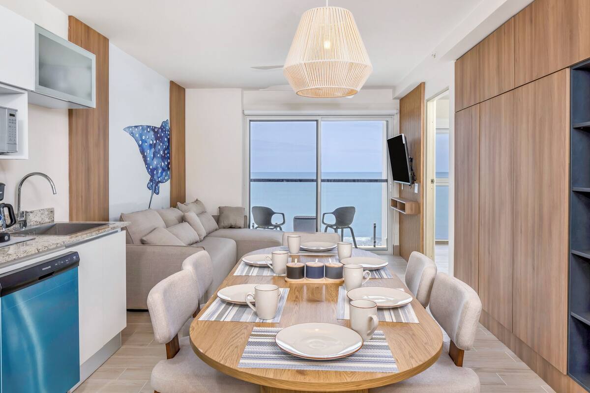 A modern kitchen with a dining area, a built-in TV, and a large window overlooking the sea.