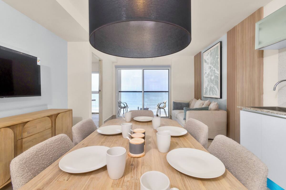 A modern dining area with a wooden table, white plates, and cups, a large black pendant light, and a view of the sea through a large window.