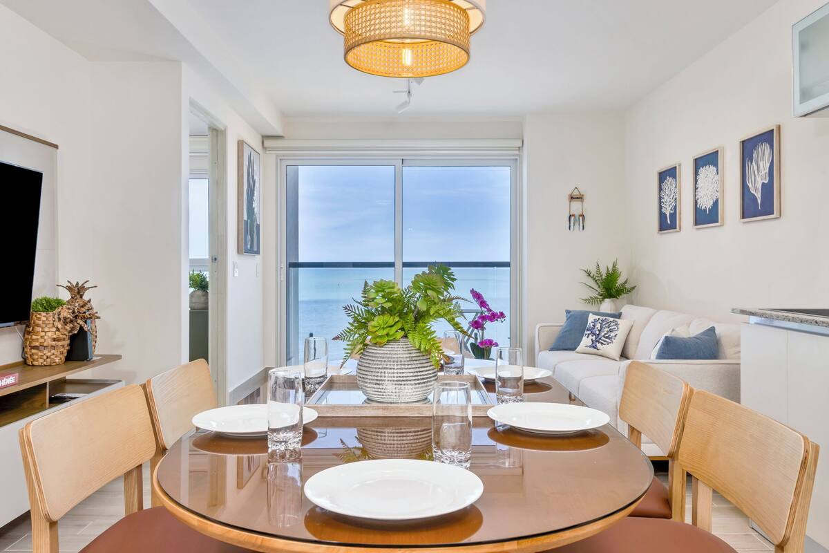 A dining area with a round table, chairs, and a view of the ocean through large windows.