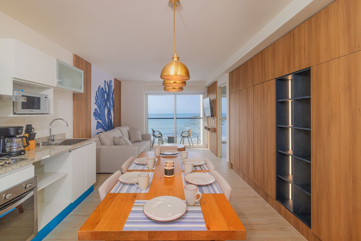 A modern kitchen with a wooden dining table, white cabinets, and a view of the sea.