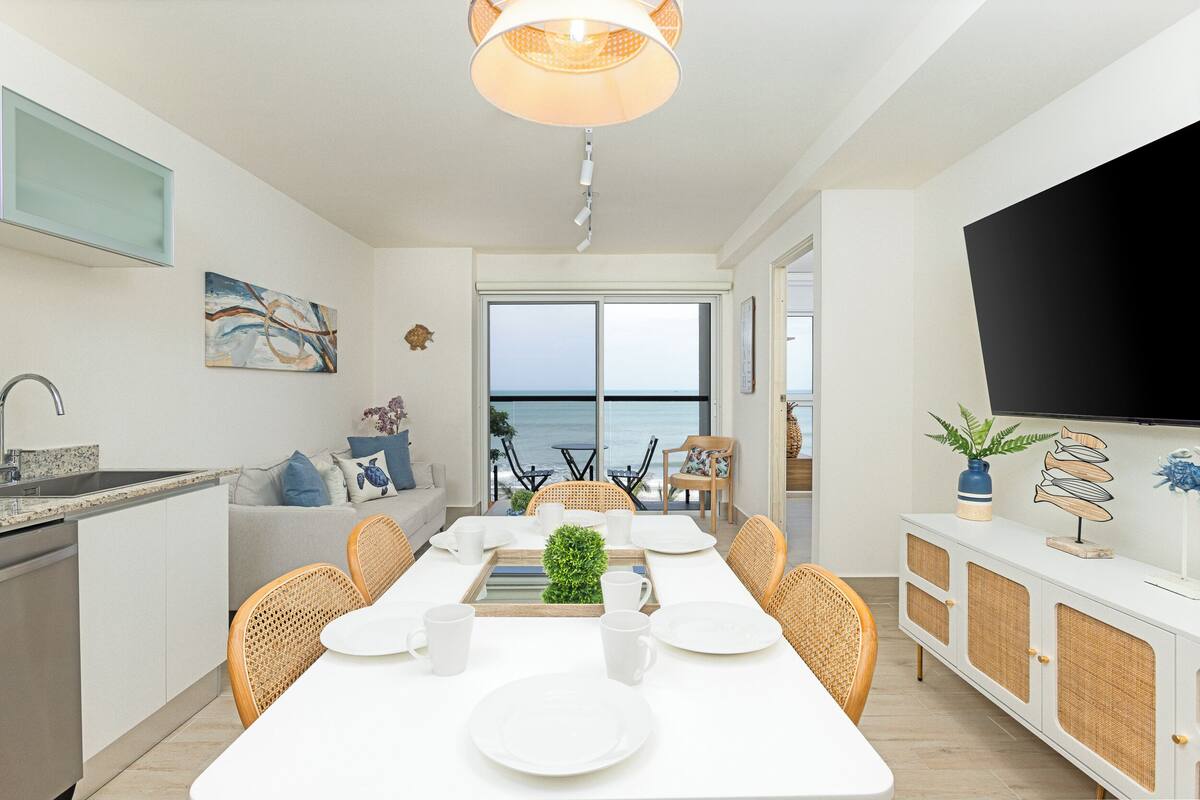 A modern kitchen with white cabinets, a dining area with a white table set for four, and a large window overlooking the ocean.