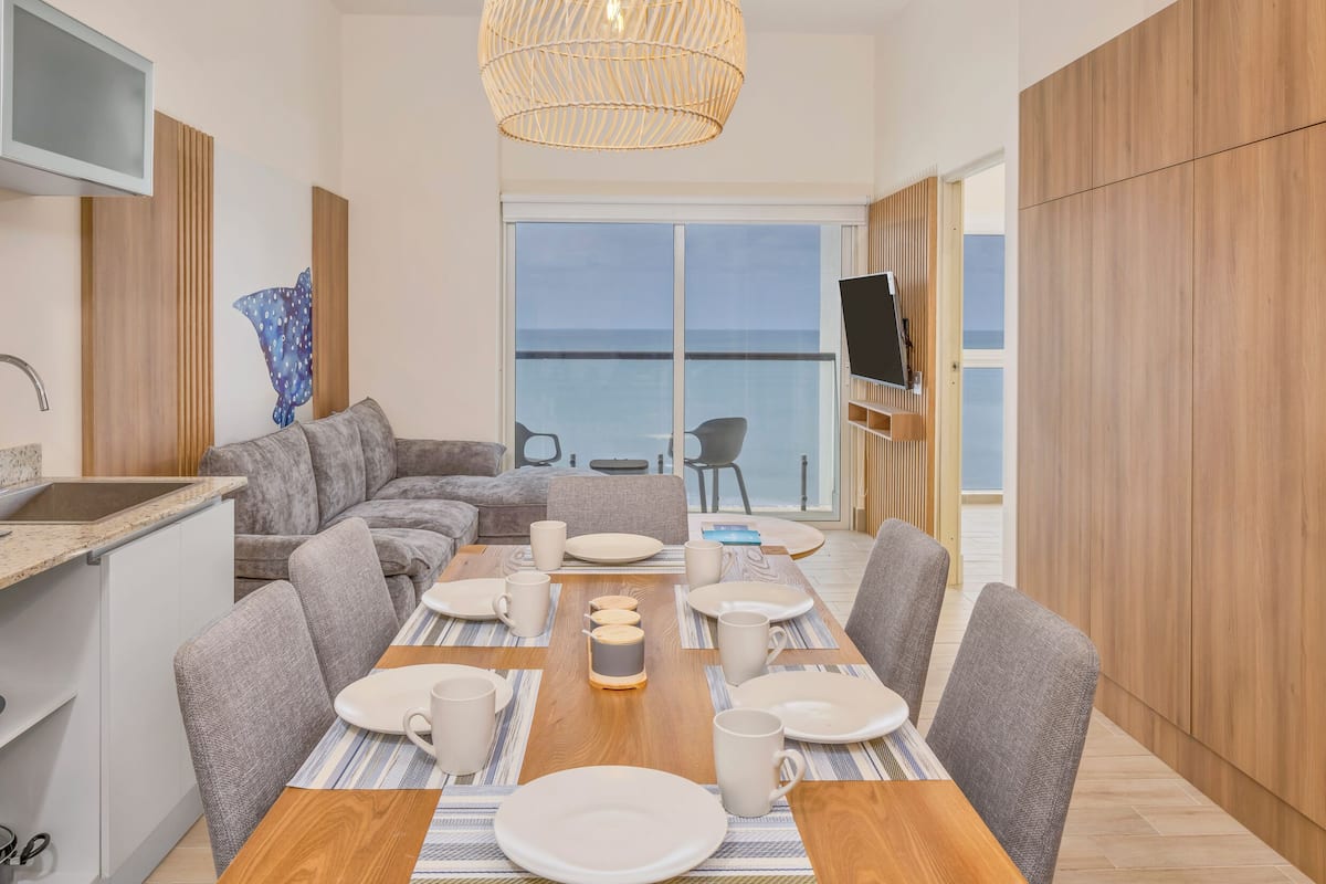 A modern dining area with a wooden table set for four, grey upholstered chairs, and a large window offering a view of the sea.