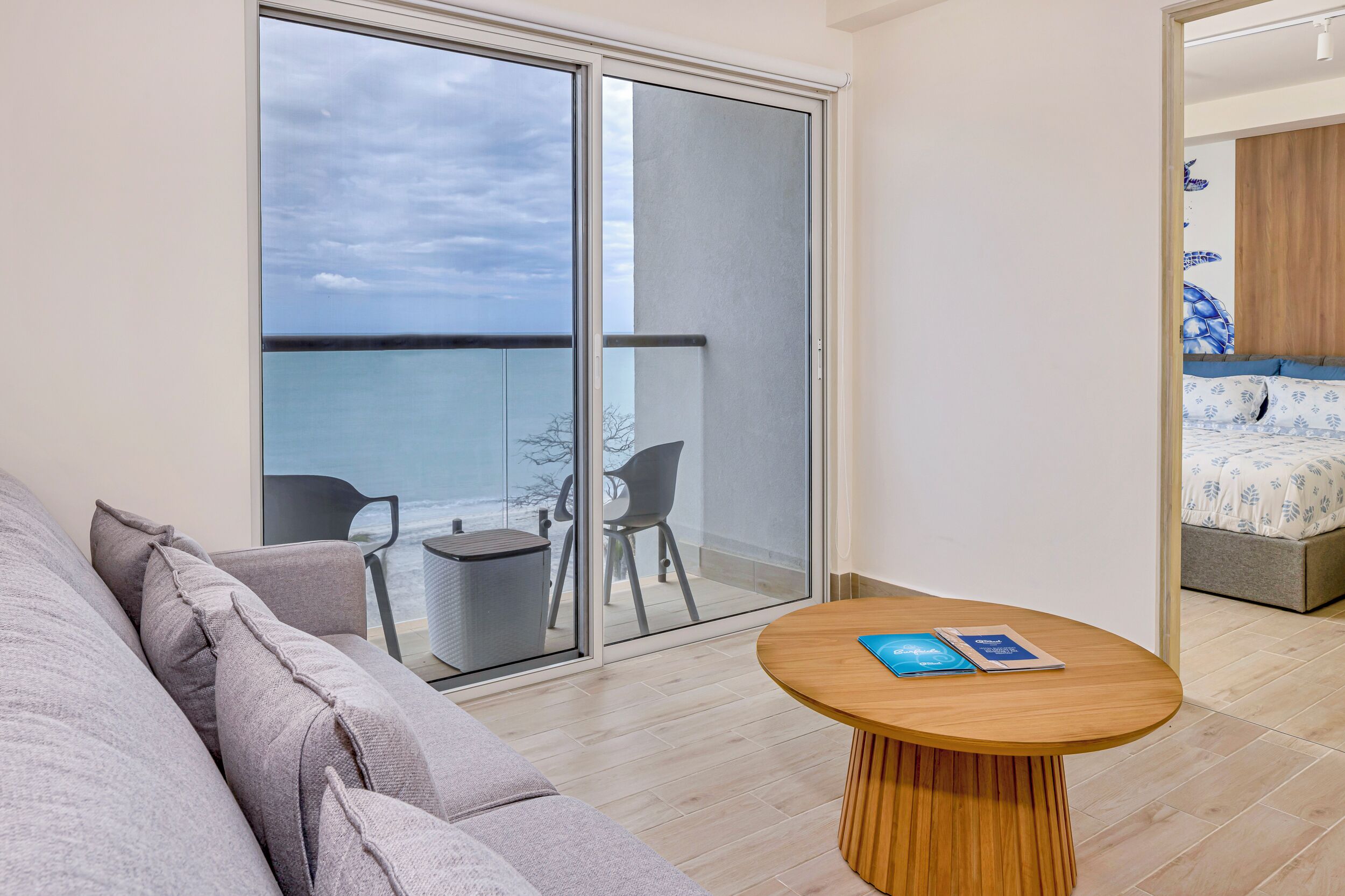 A modern room with a large sliding glass door leading to a balcony, a round wooden table, a grey sofa, and a bed with a blue and white patterned bedspread.
