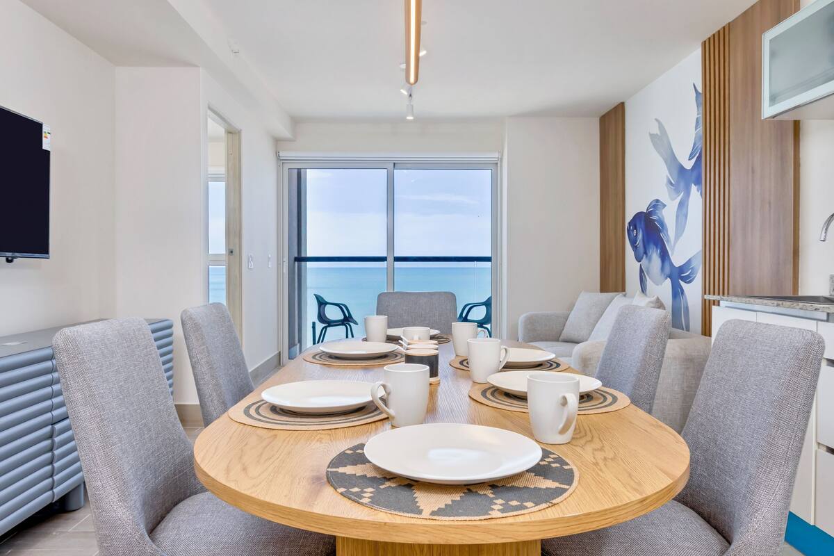 A modern dining area with a round table, grey upholstered chairs, and a view of the sea through large windows.