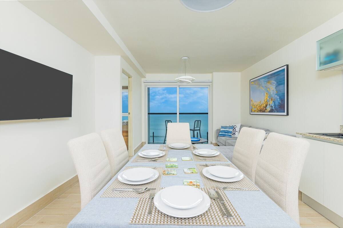 A dining area with a table set for four, a large window with a view of the sea, and a painting on the wall.
