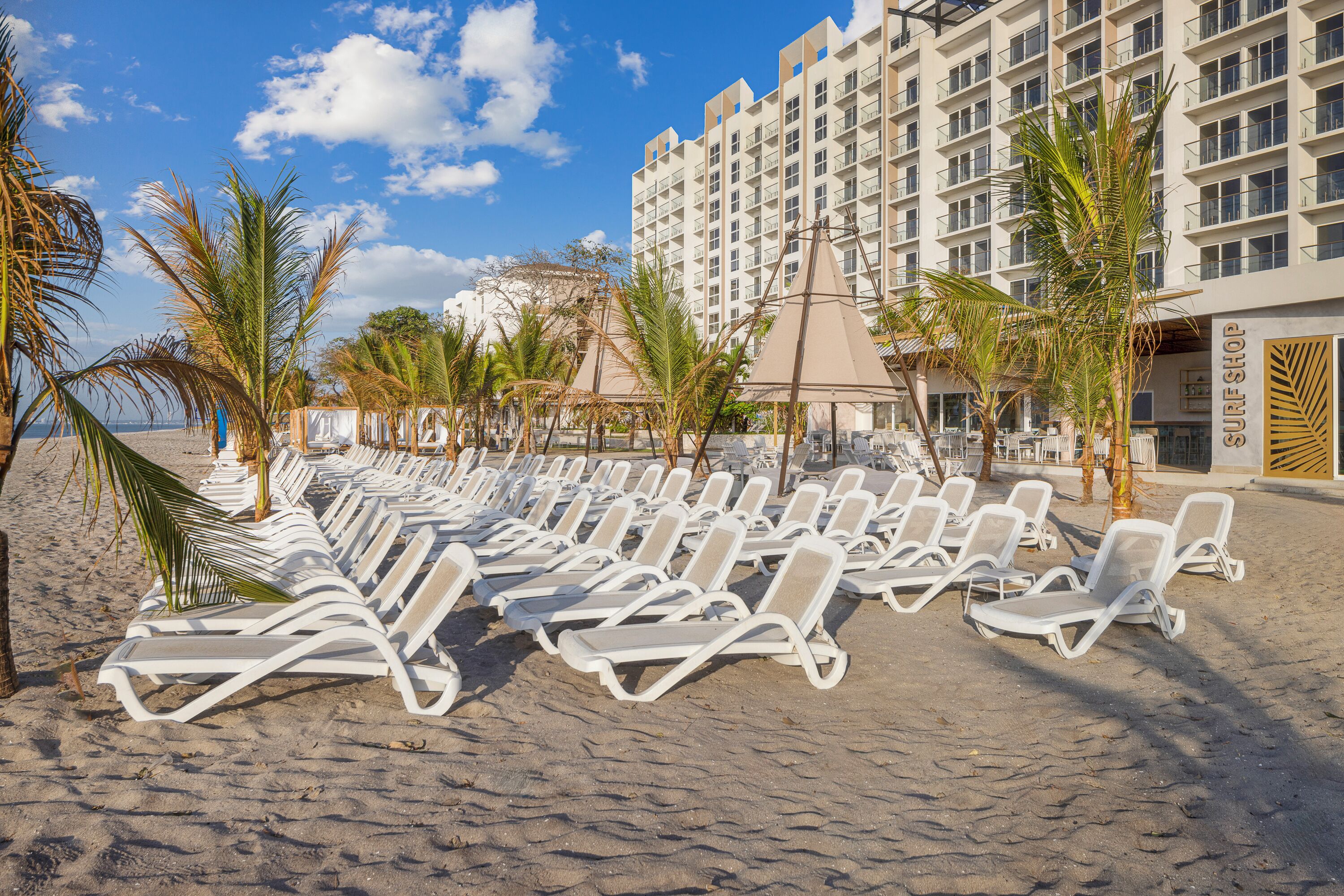 A beach with rows of white sun loungers, palm trees, and a hotel building with balconies.