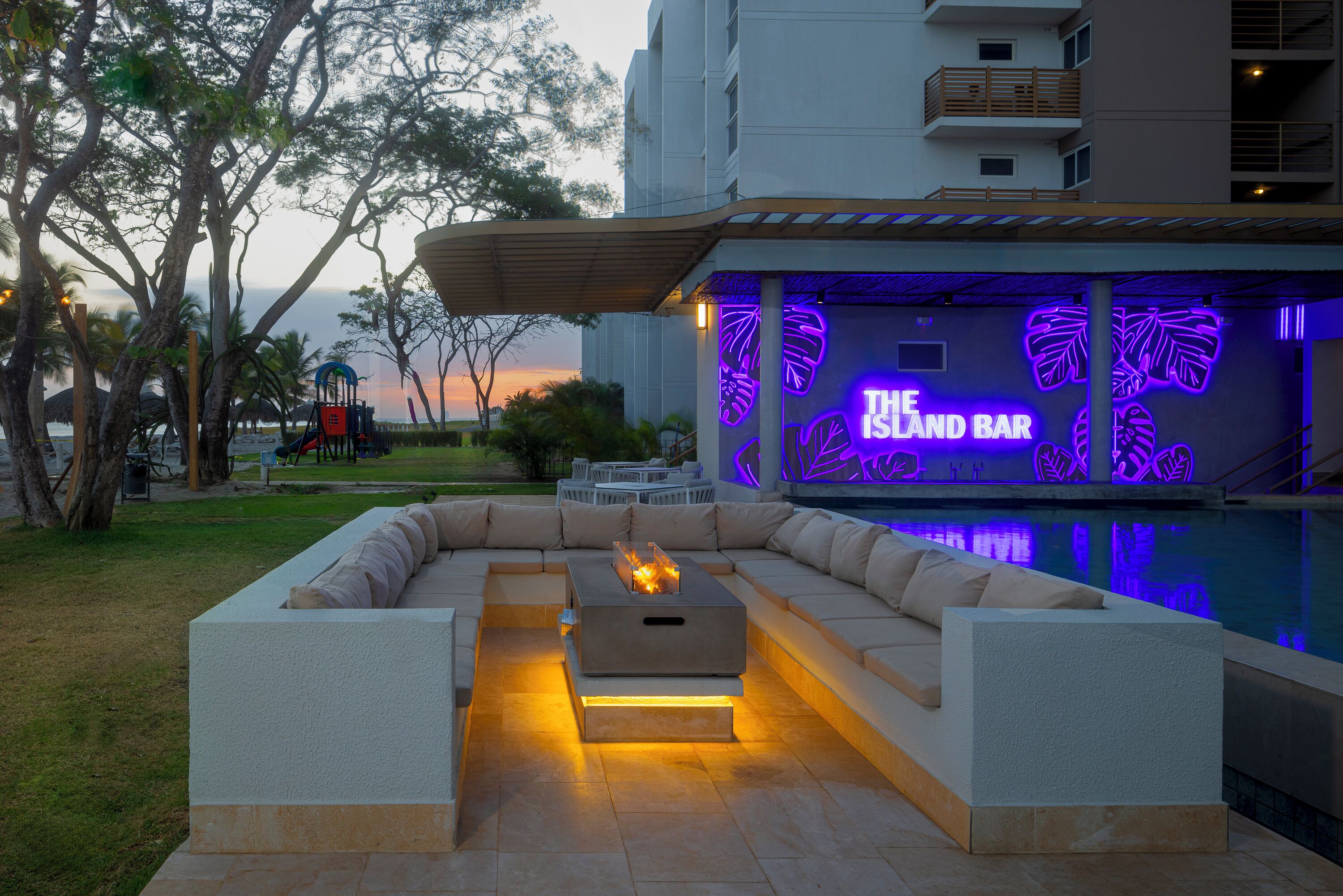 Uma área de estar externa com piscina, um bar com decorações de folhas neon e um pôr do sol ao fundo.