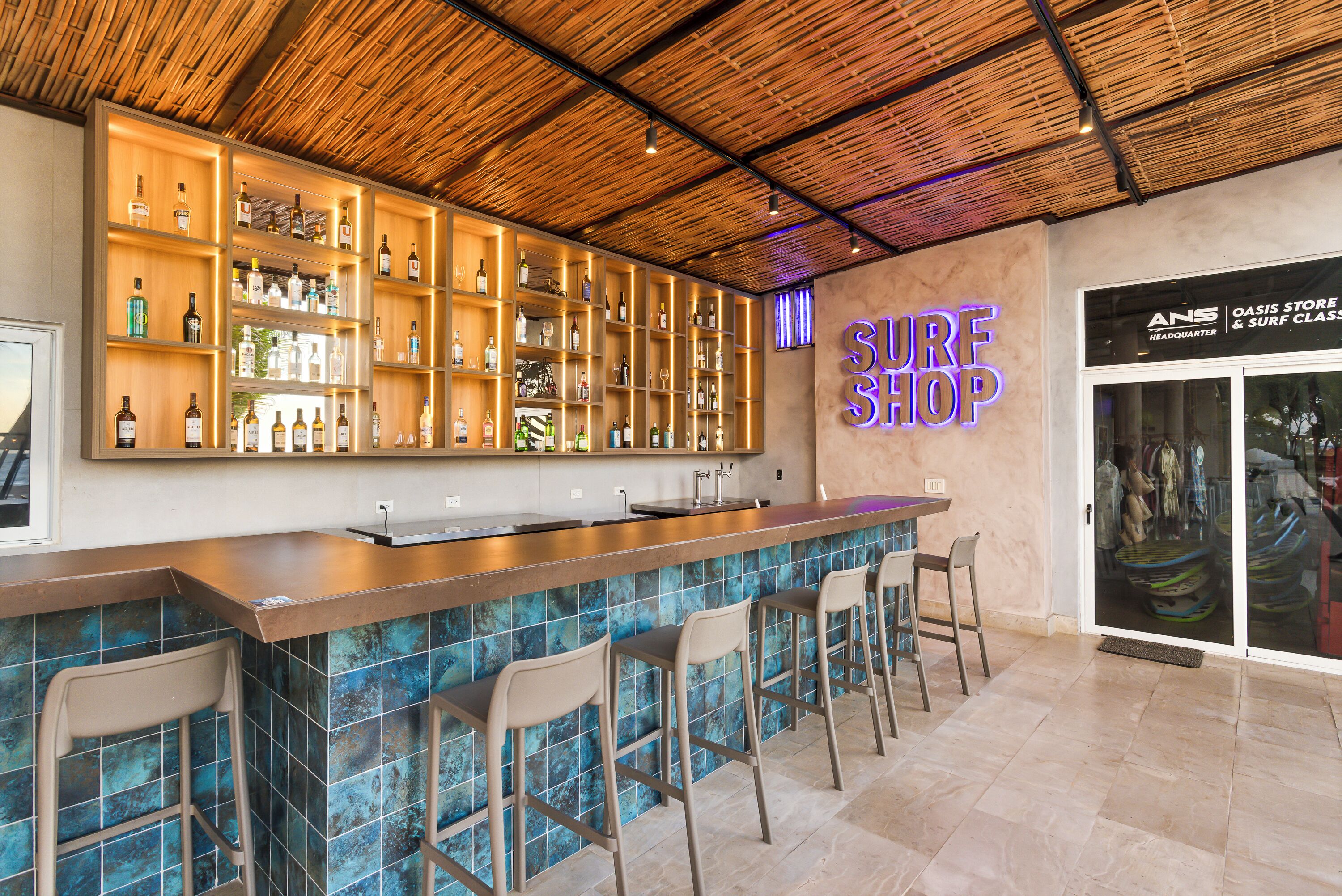 A bar area with a bamboo ceiling, shelves with bottles, and a neon sign reading 'SURF SHOP'.