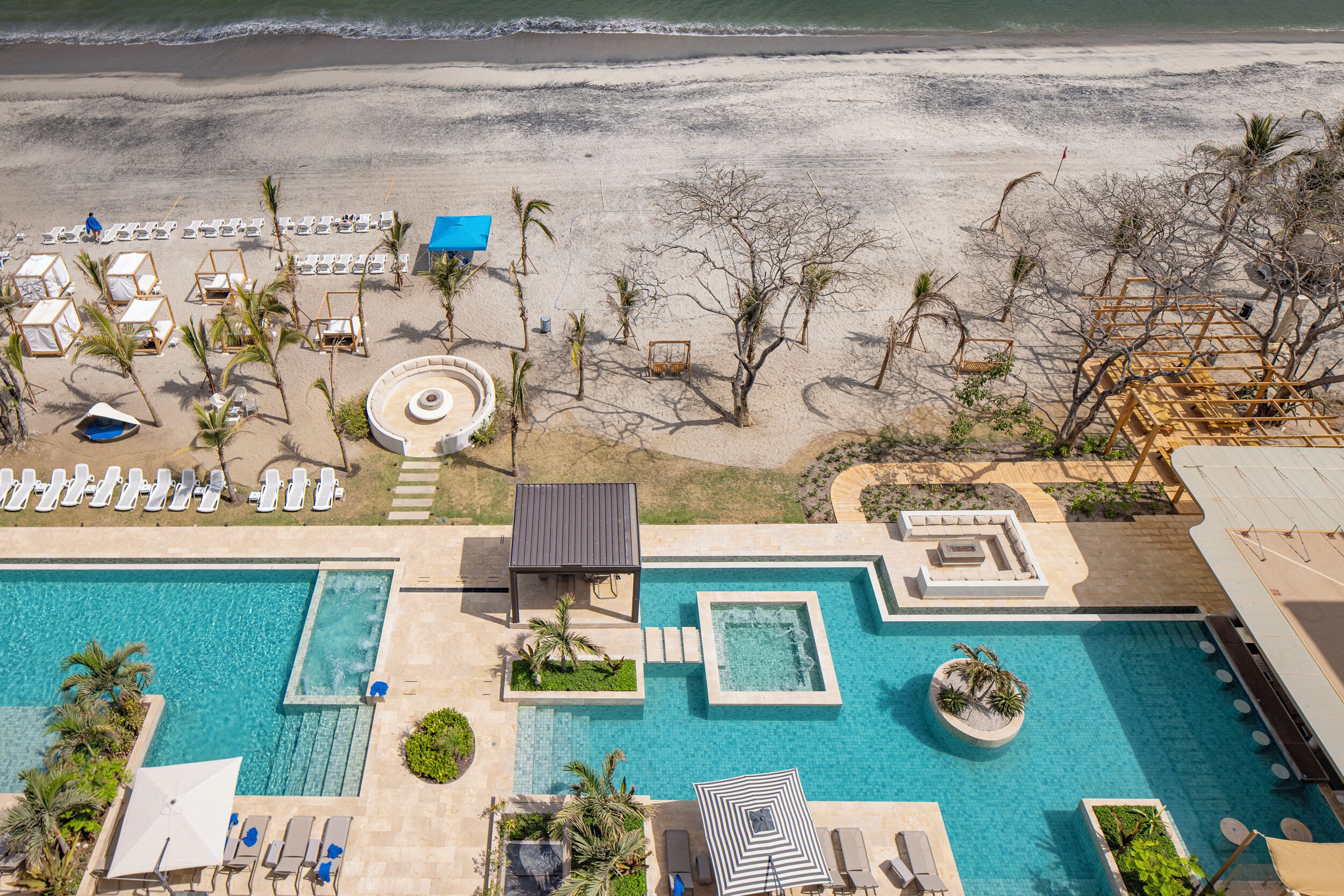 An aerial view of a resort with a large swimming pool, beachside lounge chairs, and a sandy beach.