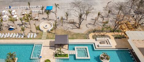 An aerial view of a resort with a large swimming pool, beachside lounge chairs, and a sandy beach.