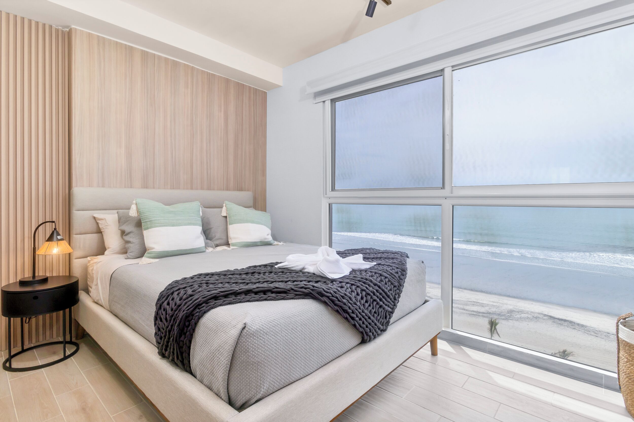 A modern bedroom with a large bed, a nightstand, and a view of the beach through a large window.