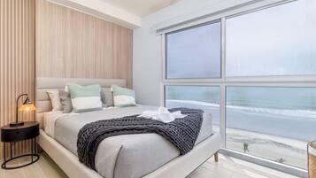 A modern bedroom with a large bed, a nightstand, and a view of the beach through a large window.