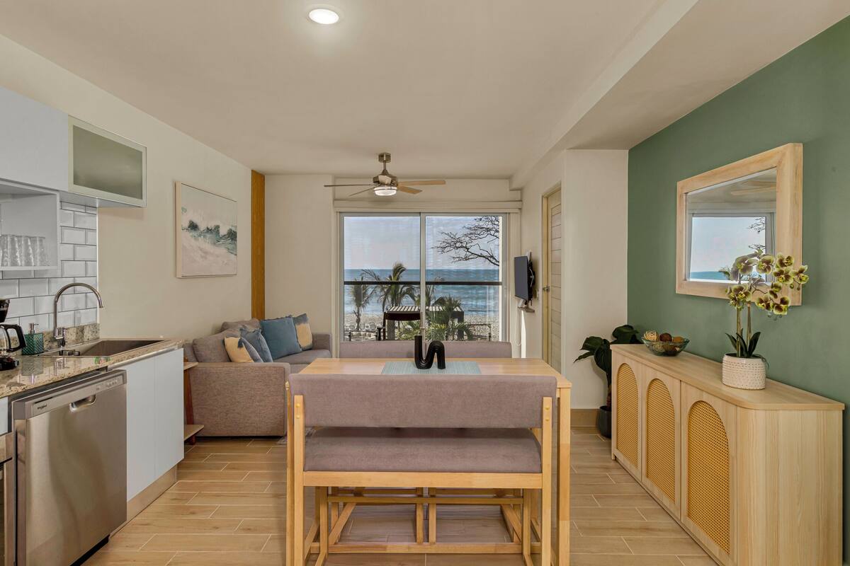 A modern kitchen with a dishwasher, a dining table with a bench, and a view of the ocean through large windows.