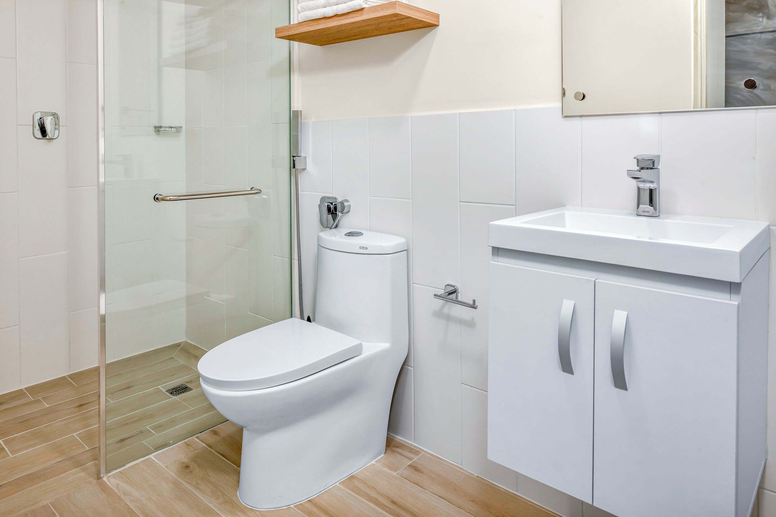 A modern bathroom with a glass shower enclosure, a white toilet, a white sink with a cabinet, and a wooden shelf above the toilet.