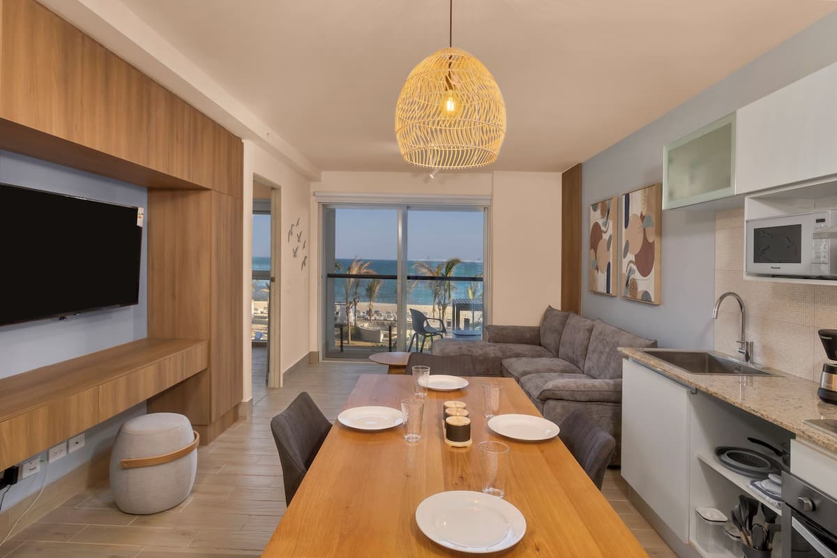 A modern living room with a wooden dining table set for four, a large flat-screen TV mounted on the wall, a cozy sofa, and a view of the beach through a large window.