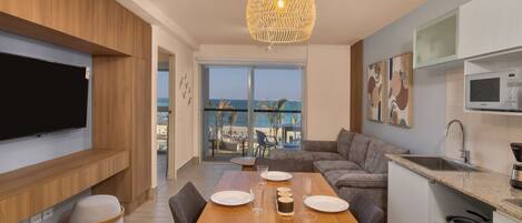 Una sala de estar moderna con una mesa de comedor de madera dispuesta para cuatro personas, un televisor de pantalla plana grande montado en la pared, un sofá acogedor y vista a la playa a través de un amplio ventanal.