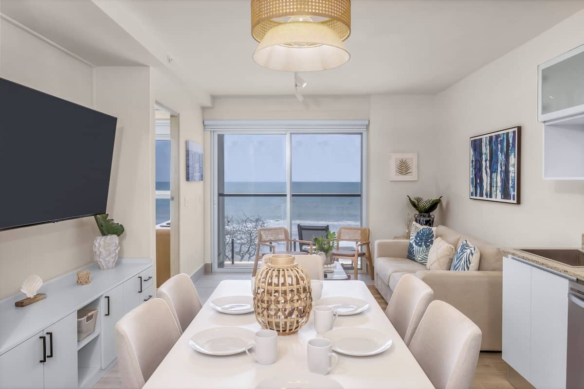 A modern dining area with a large table set for six, a television on the wall, and a view of the sea through large windows.