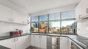 Una cocina moderna con armarios blancos, electrodomésticos de acero inoxidable, un amplio ventanal con persianas y vistas a la ciudad.