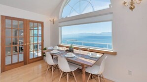 Comedor con mesa lista para cuatro, ventanas amplias con vista al mar y puerta de madera que da a otra habitación.