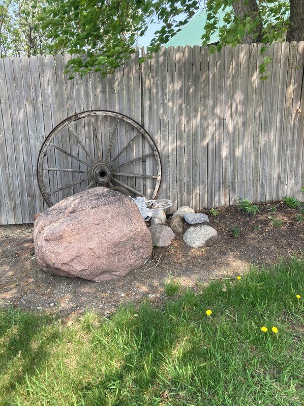A garden with a wooden fence, a large rock, a metal wheel, and some smaller stones.