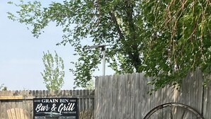A rustic outdoor seating area with wooden chairs and a large wagon wheel, a sign for 'GRAIN BIN Bar & Grill', and a wooden fence. - Rural Getaway at Homestead Lodge (Bottineau)
