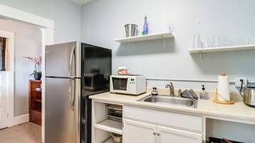 Una cocina con un refrigerador de acero inoxidable, un microondas y un lavamanos. Hay repisas sobre la encimera que contienen varios utensilios de cocina.