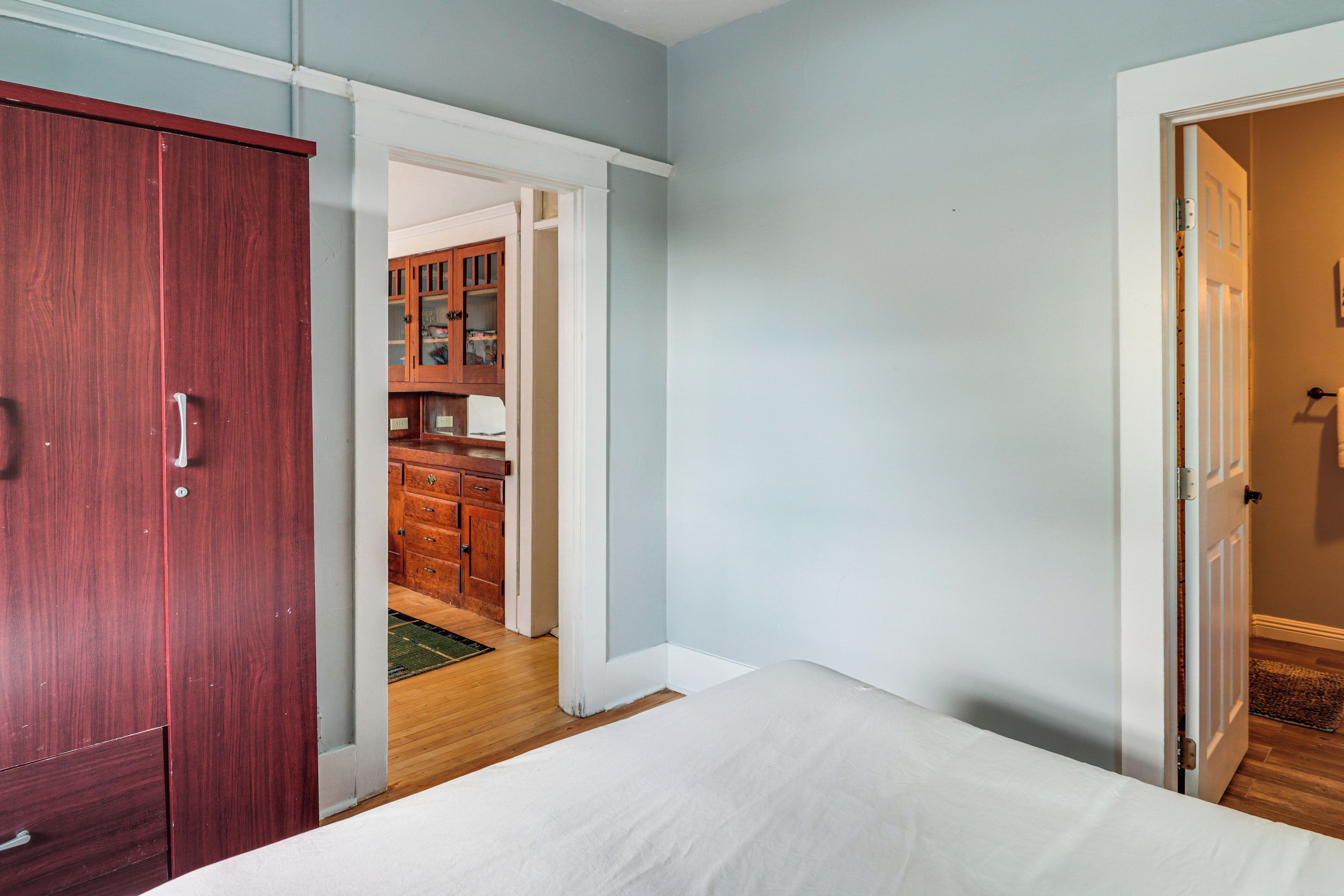 Un dormitorio con un armario de madera, una cama blanca y vistas hacia una zona de cocina con armarios de madera.