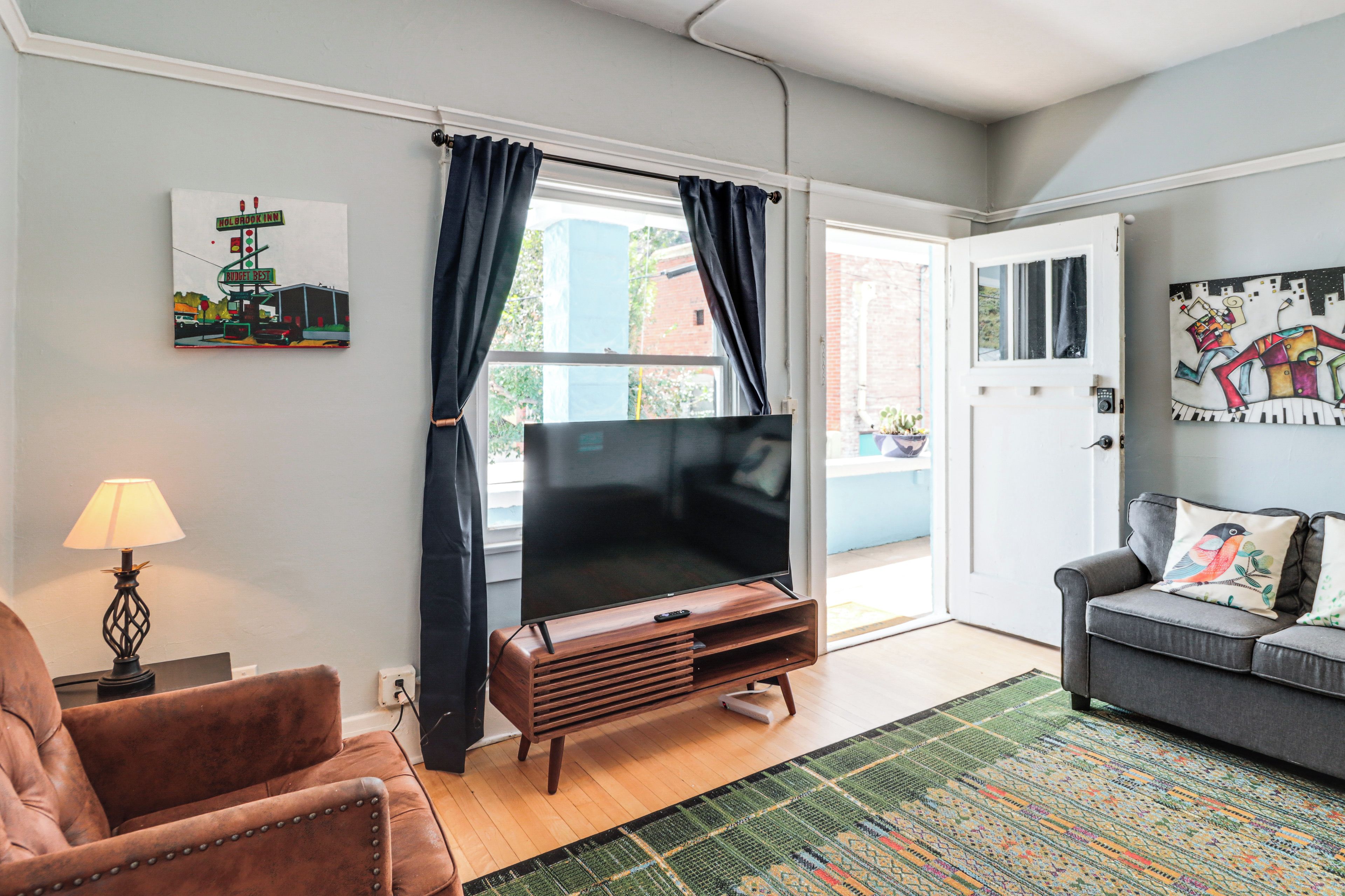 A living room with a flat-screen TV, a leather armchair, a sofa, a patterned rug, and artwork on the walls.