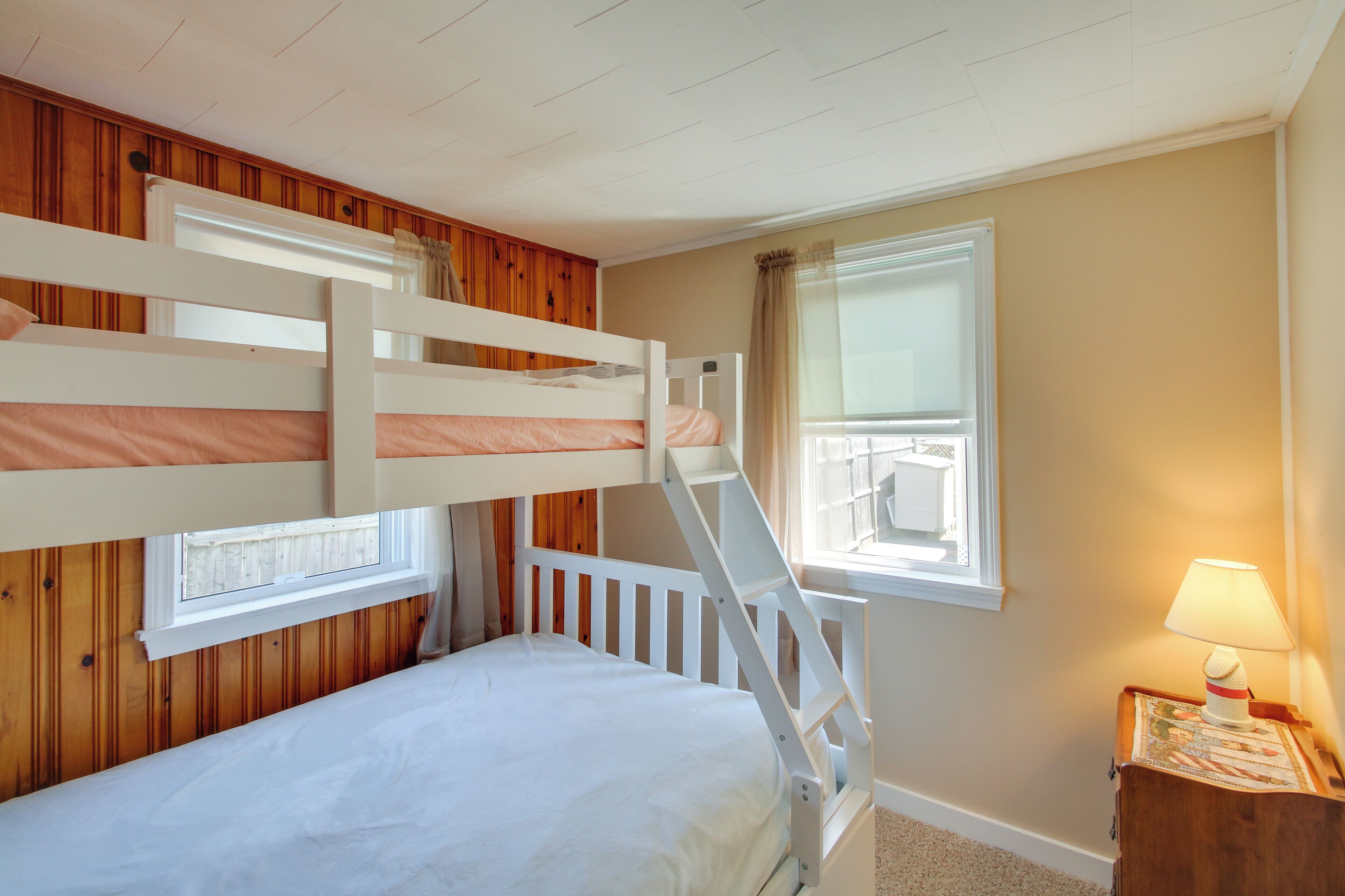 A bunk bed room with wooden paneling, a window with curtains, and a bedside lamp.