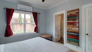 A bedroom with a bed, a window with curtains, an air conditioning unit, a door with a colorful artwork, and a small stool.