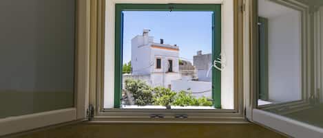 A room with a window opening to a view of buildings and a small tree.