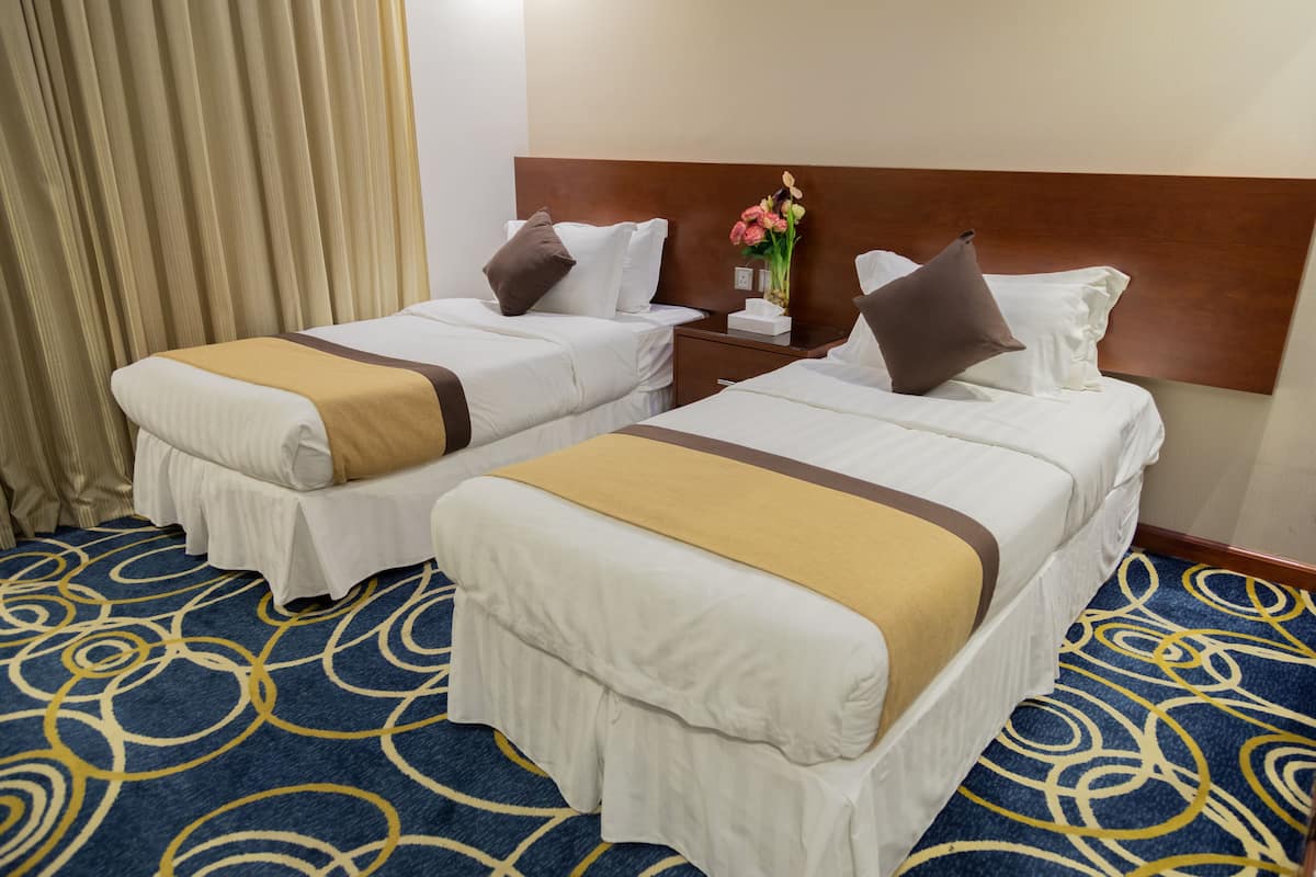 Ein Hotelzimmer mit zwei Betten, einem hölzernen Kopfteil, einem Nachttisch mit Blumenvase und einem gemusterten Teppich.