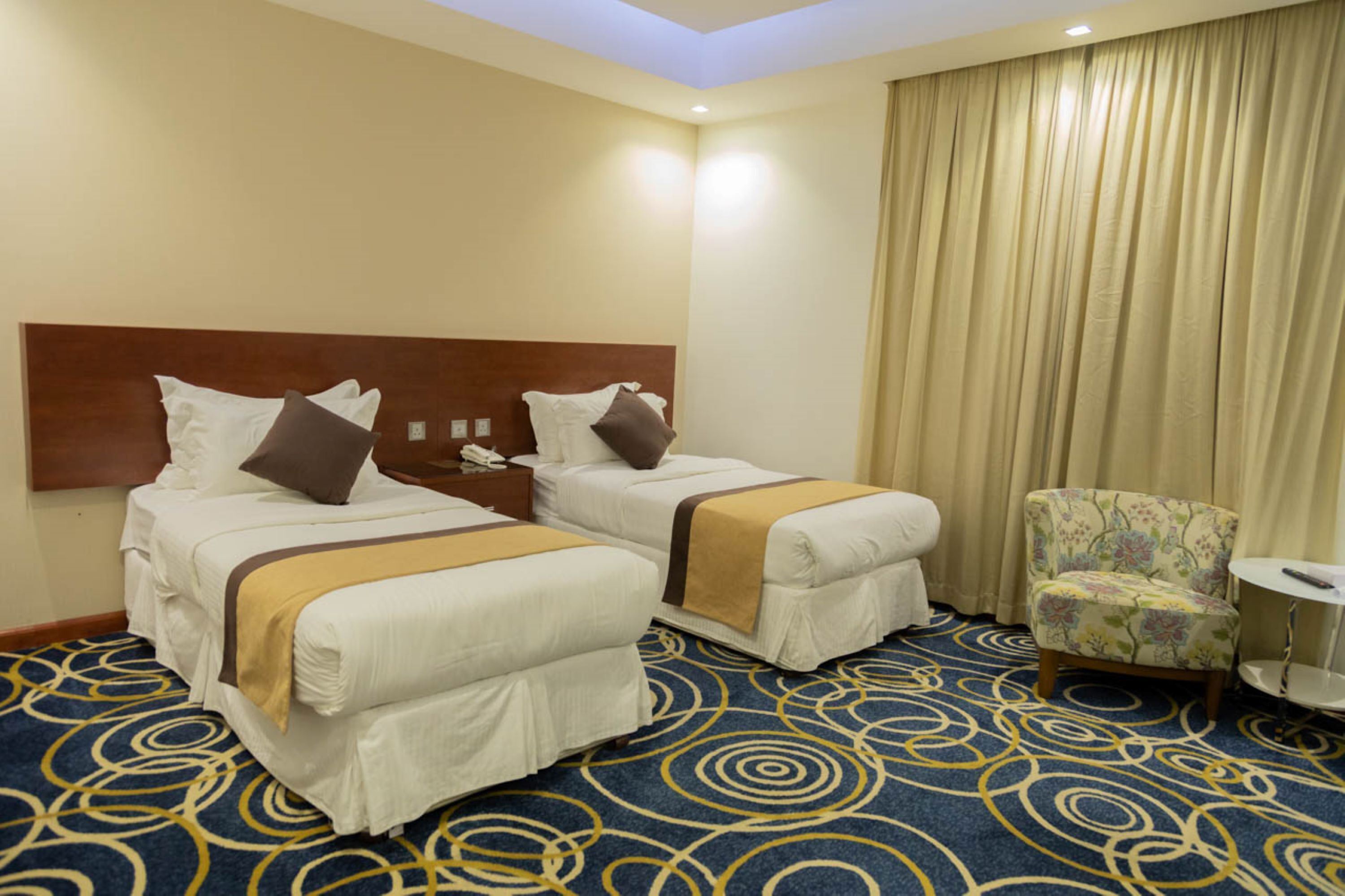 Een hotelkamer met twee bedden, een hoofdbord, een stoel, een tafeltje en een kamerbreed tapijt met een patroon.