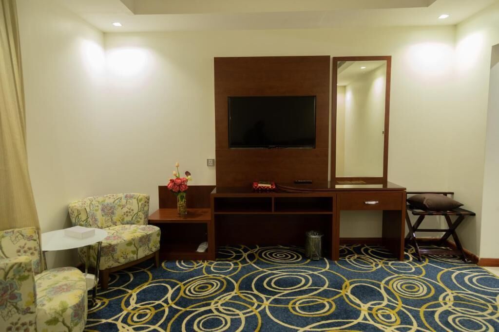 A hotel room with a television, a desk, a chair, a small table, a mirror, and a patterned carpet.