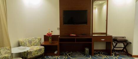 A hotel room with a television, a desk, a chair, a small table, a mirror, and a patterned carpet.