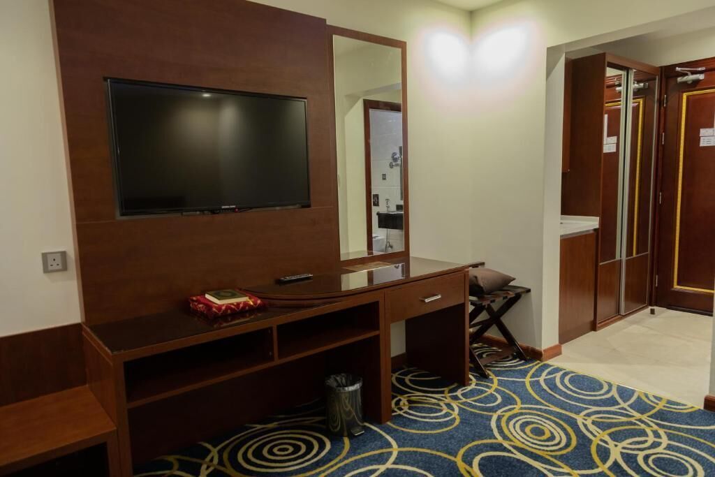 Habitación de hotel con televisor montado en la pared, escritorio con silla, espejo y armario con espejo.