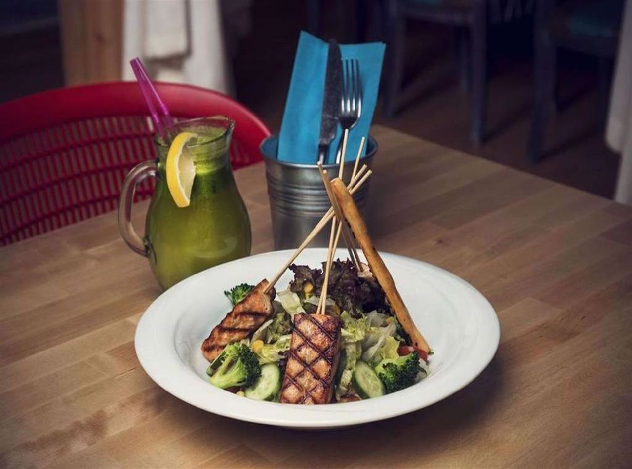 A plate of grilled chicken and vegetables on a wooden table with a pitcher of green drink and a blue napkin holder.