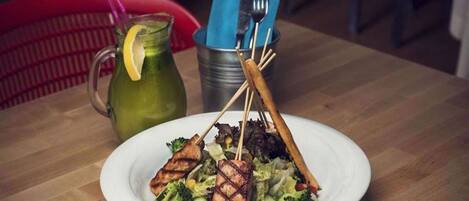 A plate of grilled chicken and vegetables on a wooden table with a pitcher of green drink and a blue napkin holder.