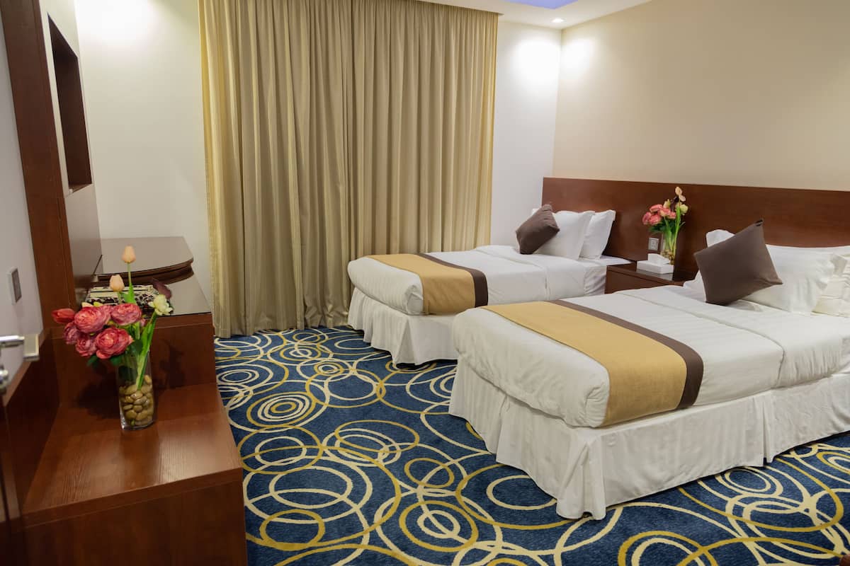 Ein Hotelzimmer mit zwei Betten, einem Holznachttisch, einer Vase mit Blumen und einem gemusterten Teppich.