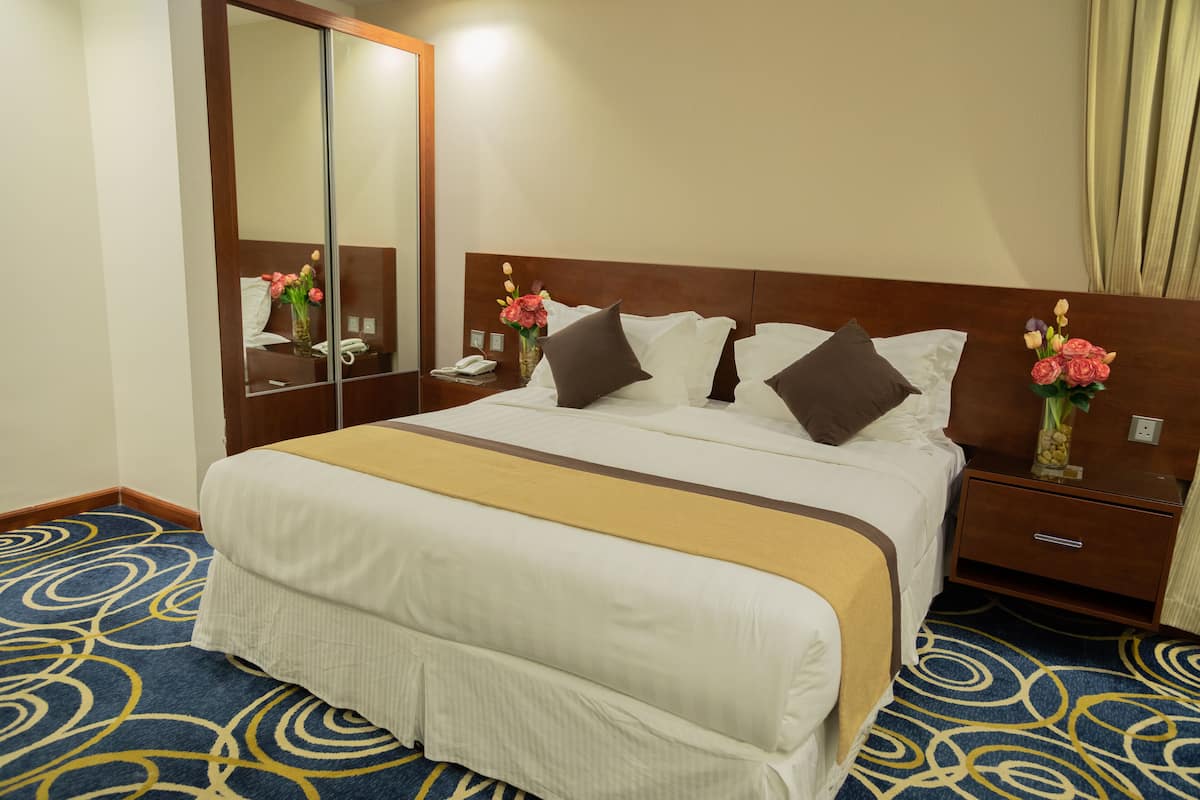 Ein Hotelzimmer mit einem großen Bett, einem Nachttisch mit Telefon, einem Spiegel und einer Vase mit Blumen.