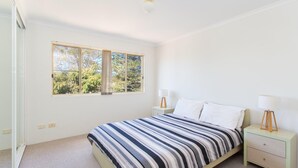 A bedroom with a large window, a bed with striped bedding, two bedside tables with lamps, and a light-colored carpet. - Haven Waters 6 - On the Lake! (Forster)