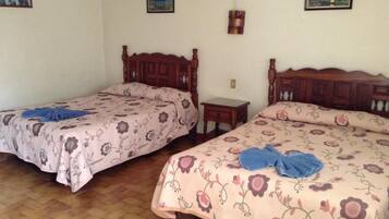 A room with two wooden beds, each with floral bedding and a blue towel on the bed. There are small bedside tables and a wall-mounted lamp.