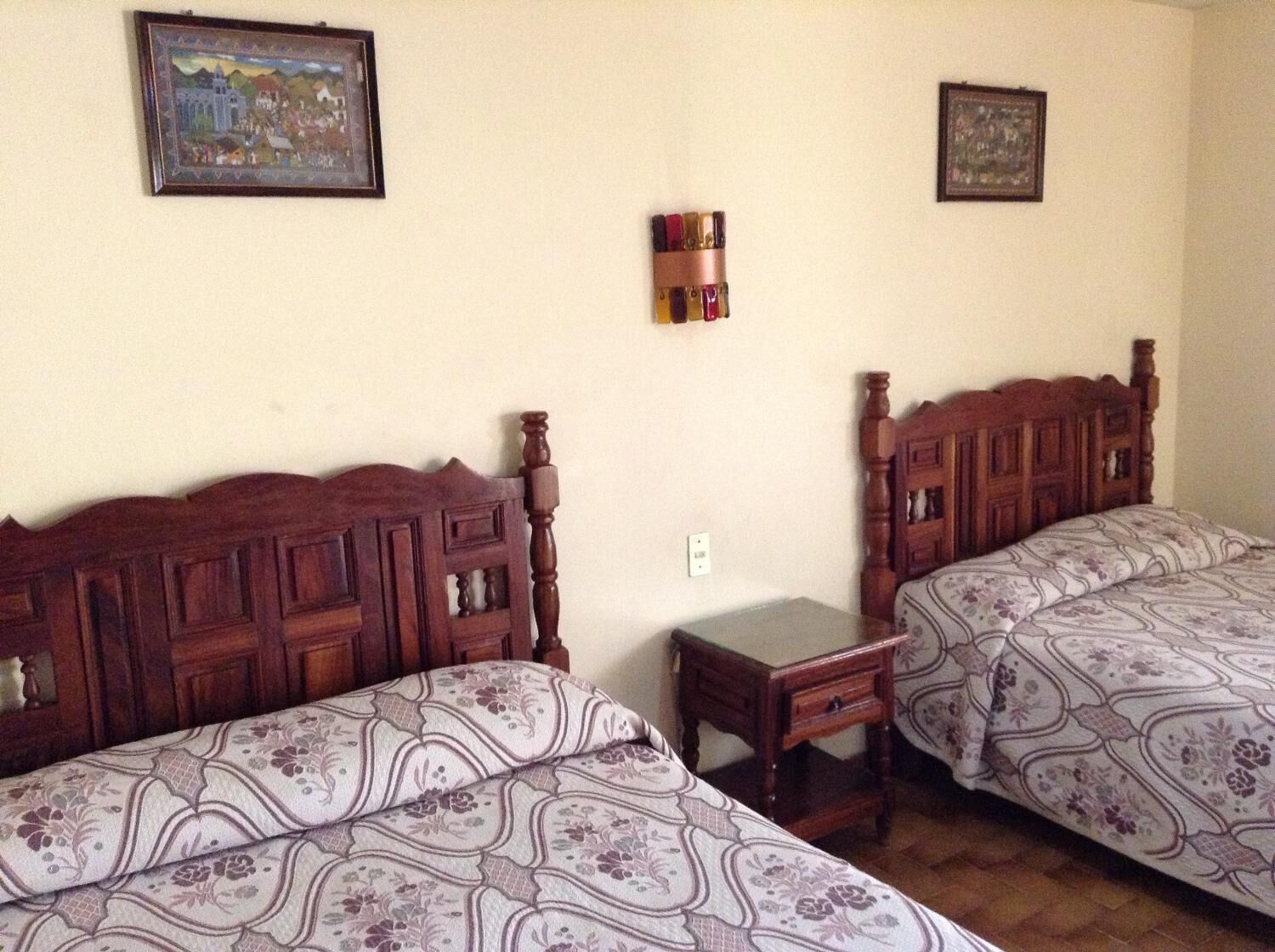 Una habitación con dos camas de madera, ropa de cama floral, una mesita de noche pequeña y una lámpara fijada a la pared.