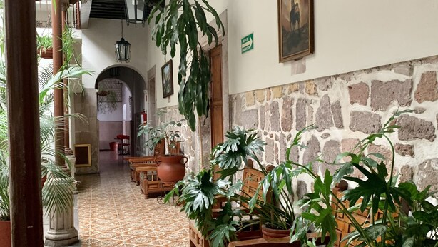 A corridor with patterned tile flooring, wooden furniture, and potted plants. - Hotel Atilanos (Morelia)